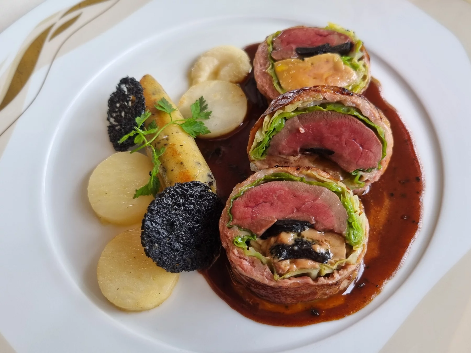 Tournedos de pigeon emballé dans une crépine de veau, farci au foie gras d’oie, à la truffe noire et au chou, servi avec une sauce périgourdine et une déclinaison de céleri, en gnocchi, rôti et en purée.